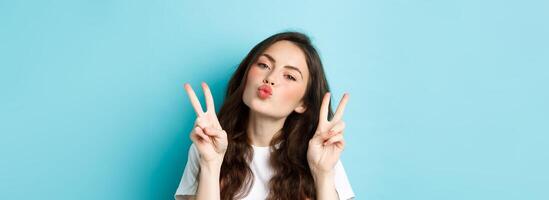 Close up portrait of glamour brunette girl making kissing face, showing kawaii v-sign and smiling cute at camera, standing happy against blue background photo