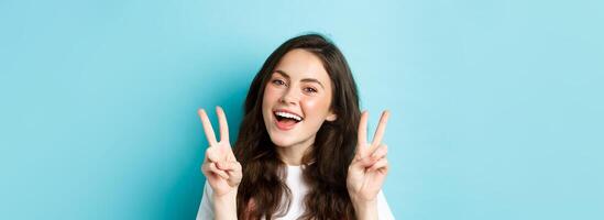 Young positive modern girl sending good vibes only, showing v-sign and smiling happy, standing against blue background photo