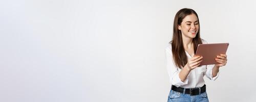 Portrait of beautiful girl smiling, looking happy, watching on digital tablet, standing against white background photo