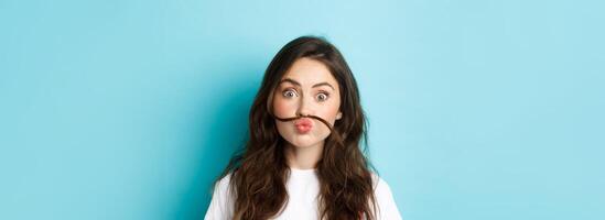 Close up portrait of funny silly girl playing around, making moustache with hair strand above lips, standing over blue background photo
