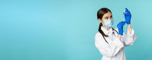 Vaccination from covid and healthcare concept. Young woman doctor, nurse in face mask and gloves, using syringe to do vaccine shot, standing over torquoise background photo
