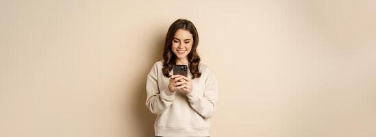 People and cellular technology. Beautiful stylish woman using mobile phone, smartphone app, smiling and looking at screen, standing over beige background photo