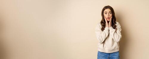 Image of surprised, amazed brunette girl gasping, looking fascinated at camera, standing over beige background photo