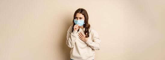 Covid-19 and health concept. Sick woman in medical face mask coughing, feeling ill with sour throat, standing over beige background photo