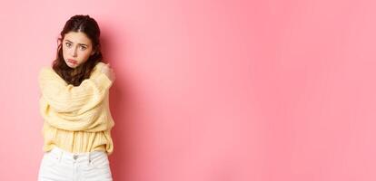 Cute silly girl making sad face and looking with puppy eyes at camera, hugging herself, standing innocent against pink background photo