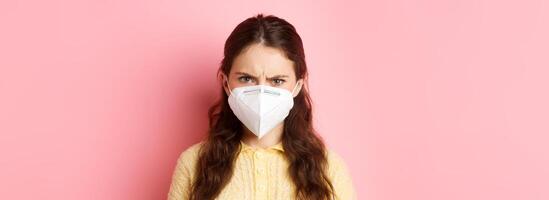 Preventive measures, health care concept. Close up of angry woman in respirator frowning, looking with judgemental face at person without mask, standing against pink background photo