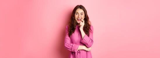 Beautiful excited girl smiling, biting finger and looking with tempted face at camera, having interesting idea, looking at something intriguing, standing against pink background photo