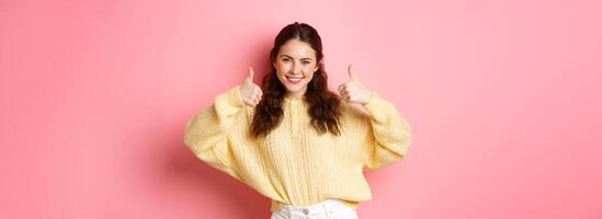 Well done, very good. Smiling young woman showing thumbs up in approval, like your choice, praise good thing, standing satisfied against pink background photo