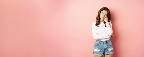 Image of sad and bored, reluctant young woman looking at smth boring, feeling lonely, leaning head on fist with indifferent face expression, standing over pink background photo