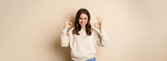 Okay. Confident smiling modern girl showing ok sign, approve, like and recommend smth good, standing over beige background photo