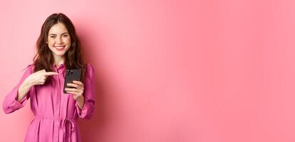 Pretty caucasian girl advertising online offer, pointing finger at her smartphone and smiling satisfied, recommending app, standing over pink background photo