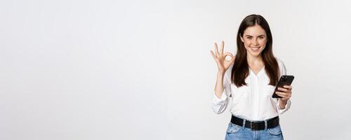 Smiling woman showing okay, ok sign while using mobile phone, approve app on smartphone, excellent service, online store, standing over white background photo