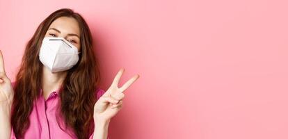 Coronavirus, preventive measures and health concept. Close up portrait positive woman in medical respirator, posing and looking happy with peace v-signs, pink background photo