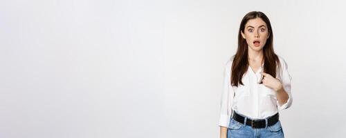 Portrait of young woman pointing at herself with disbelief, surprised face expression, being named or chosen, standing over white background photo