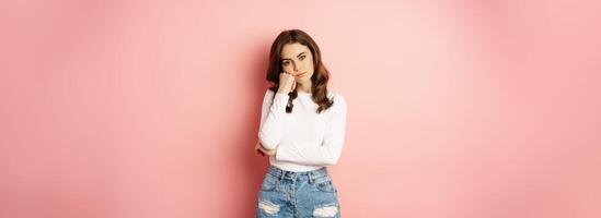 Image of sad and bored, reluctant young woman looking at smth boring, feeling lonely, leaning head on fist with indifferent face expression, standing over pink background photo