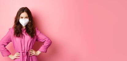 Covid-19, pandemic and lifestyle concept. Angry woman in medical respirator look with judgement and dislike, frowning and scolding someone without face mask, pink background photo