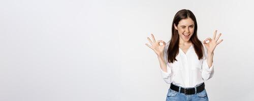Smiling confident business woman, winking, showing okay ok sign, excellent, no problem gesture, standing over white background photo