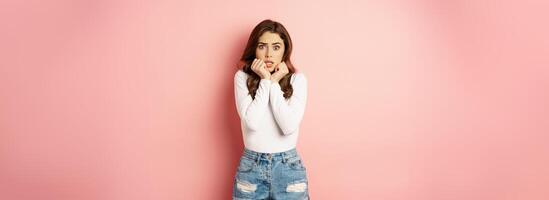 Portrait of scared brunette girl, shaking from fear, looking afraid at camera, biting fingernails, standing against pink background photo
