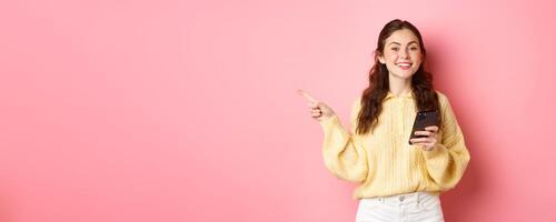 Beautiful modern girl holding smartphone and pointing finger aside, showing promotional text on copyspace while using mobile phone, standing over pink background photo