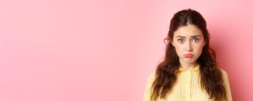 Close up of cute and coy girl making sad pouting face to ask for something, wants pity, standing against pink background gloomy photo