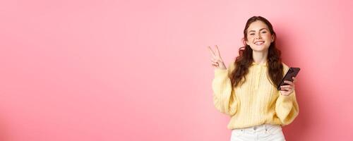 Technology and online shopping. Happy young woman smiling pleased, holding smartphone, showing v-sign and look positive at camera, standing over pink background photo