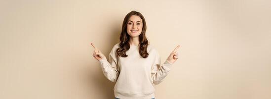 Cheerful caucasian woman showing directions, two ways, pointing sideways at variants, choices for customer, standing over beige background photo