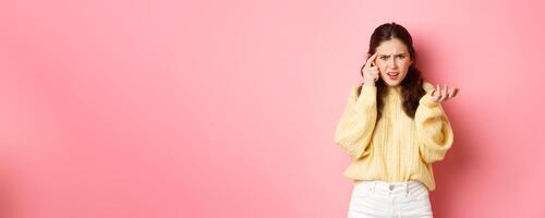 Confused and annoyed girl pointing at head with index finger, scolding someone for stupid or crazy behaviour, being disappointed, standing against pink background photo