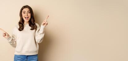 Cheerful caucasian woman showing directions, two ways, pointing sideways at variants, choices for customer, standing over beige background photo
