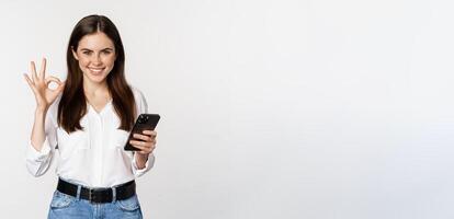 Smiling woman showing okay, ok sign while using mobile phone, approve app on smartphone, excellent service, online store, standing over white background photo