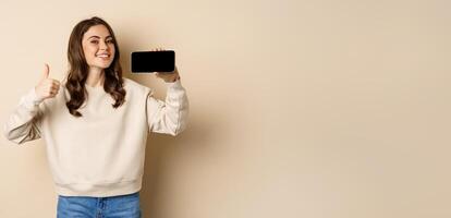 Smiling happy woman showing smartphone horizontal screen, thumbs up, recommending website, online store or app, standing over beige background photo