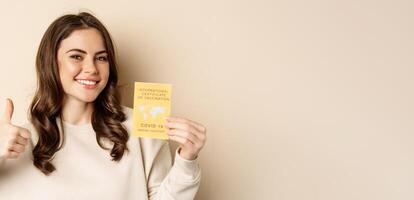 Covid-19 vaccination concept. Portrait of happy healthy girl showing coronavirus international vaccination certificate and thumb up, travelling during pandemic, beige background photo