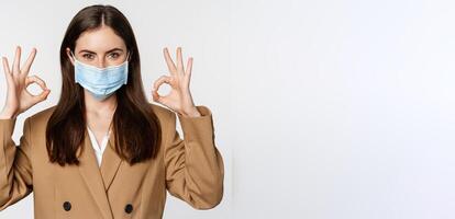 Workaplce and pandemic concept. Close up portrait of business woman showing okay signs, wearing medical mask, recommend, praise something, standing over white background photo