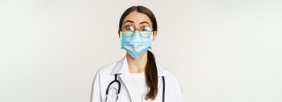 Medical worker, young doctor looking amazed and surprised, wearing face mask to prevent catching covid-19, standing over white background photo