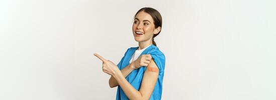 Smiling nurse, female healthcare worker in scrubs, pointing finger right, showing patch on shoulder, vaccination campaign, covid-19 and pandemic concept, white background photo