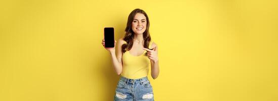 Young stylish woman showing mobile phone screen, pointing at smartphone display, app interface or online store, smiling pleased, standing over yellow background photo