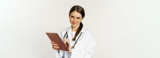 Doctor in white coat using digital tablet, reading medical data on gadget, working in hospital, standing over white background photo