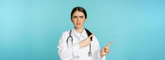 Confused, disappointed medical worker, frowning while pointing fingers left with doubtful, puzzled face expression, standing over torquoise background photo