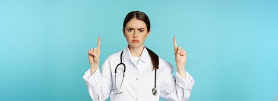 Sad and disappointed healthcare worker, doctor pointing fingers up, frowning upset, showing bad news, standing over torquoise background photo