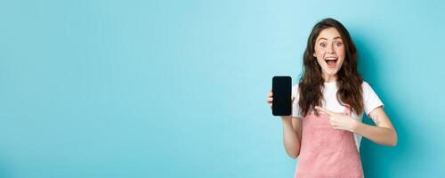 Wow check this out. Excited pretty girl pointing finger at phone screen, showing logo or store advertisement on smartphone, standing against blue background photo