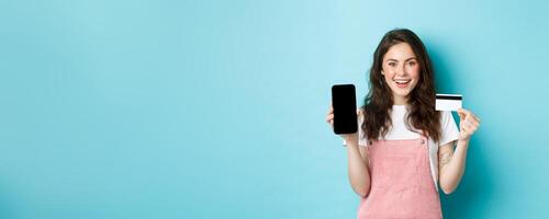 Image of beautiful smiling woman in stylish outfit, showing plastic credit card and empty smartphone screen, recommend app, standing over blue background photo
