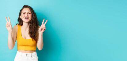 Summer holidays and emotions concept. Happy beautiful girl showing peace v-signs and having fun, laughing and smiling at camera, standing against blue background photo