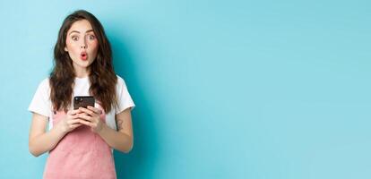 Portrait of brunette cute girl look surprised after using smartphone, holding mobile phone and say wow at camera with impressed face, blue background photo