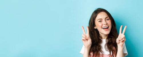 Young positive modern girl sending good vibes only, showing v-sign and smiling happy, standing against blue background photo