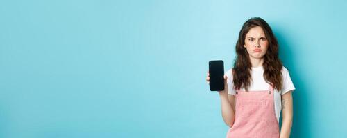Disappointed girl frowning while showing empty smartphone screen, look displeased or angry, standing against blue background photo