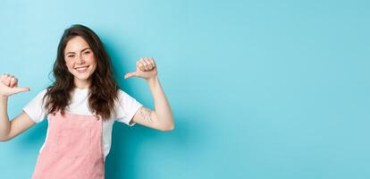 Young smiling woman pointing at herself, looking confident and happy, self-promoting, talking about her, personal achievement, standing over blue background photo
