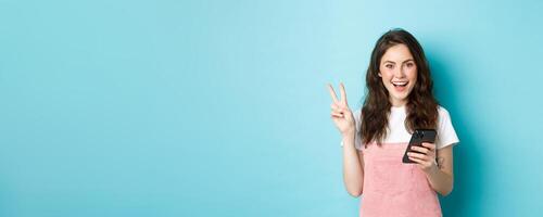 Online shopping. Positive smiling girl showing v-sign and looking happy at camera, using smartphone, holding mobile phone, standing against blue background photo