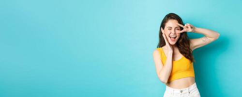 Excited girl screaming from joy, ready for summer holidays, showing v-sign and smiling happy, standing happy against blue background photo
