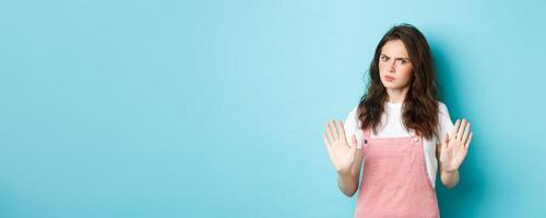 Stop right there. Serious young woman frowning, raising hands and showing block, say no, disapprove bad thing, rejecting you, standing against blue background photo