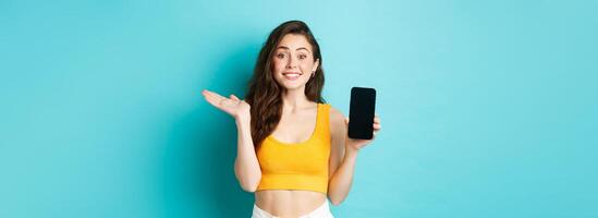 Cheerful young girl introduce your app or store, showing emtpy smartphone screen and smiling at camera, standing against blue background photo