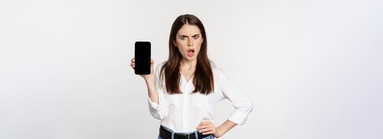 Shocked and frustrated woman showing mobile phone screen app, smartphone application, looking insulted, standing over white background photo